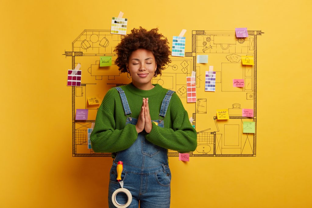 Calm African American woman prays for good result of work, makes repair of home, poses against house design project, busy doing repairs, dressed casually, has screwdriver and sticky tape in pocket